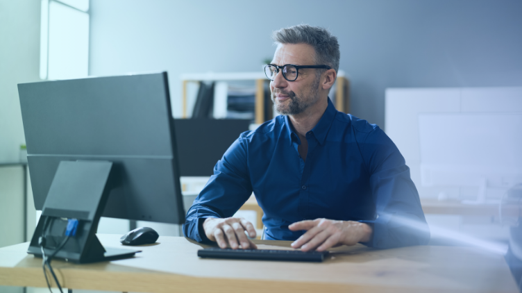 Middle-aged man wearing glasses and a blue shirt working on a desktop computer at his desk in an office. 